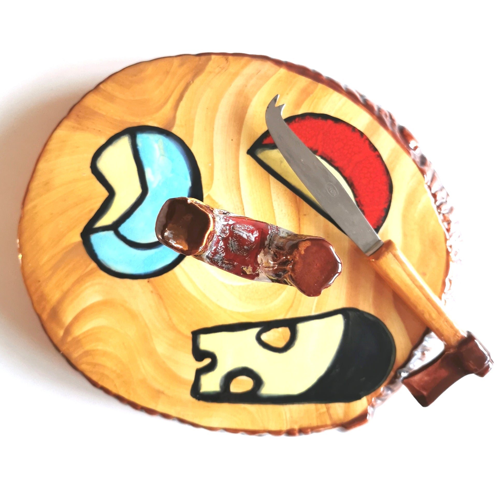Plateau à fromage vintage en céramique trompe-l'œil imitant une section de bûche de bois. Le décor central présente des motifs de fromages stylisés (bleu, rouge, emmental) sur un fond jaune veiné. Le manche du plat imite un sarment de vigne et le couteau assorti possède un manche en forme de hache miniature. Style kitsch rustique, quelques éclats d'émail visibles."